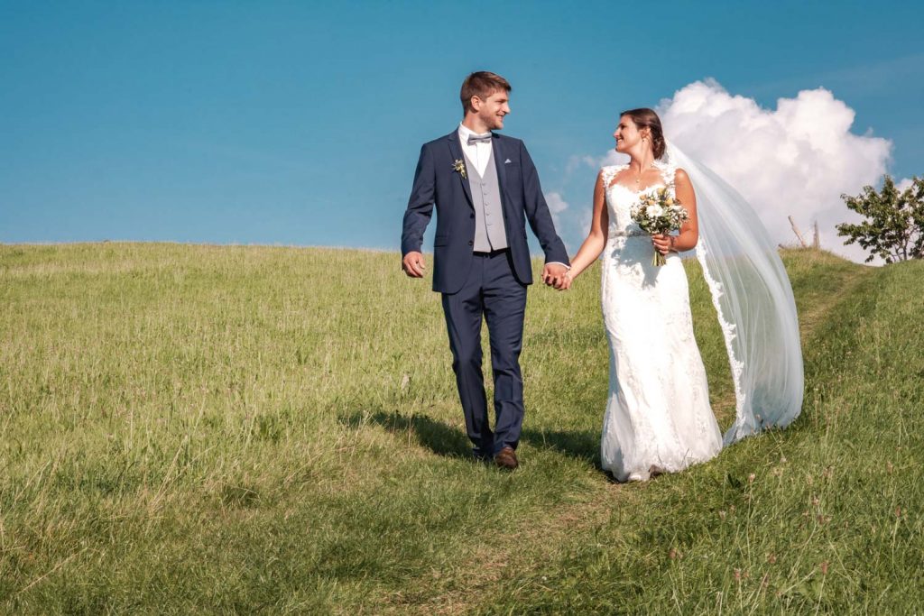 Hand in Hand geht das Brautpaar nach der Trauung in Schliersee über den Irschenberg bei traumhaftem Sommerwetter
