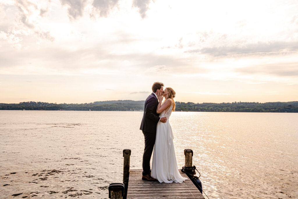 Glücklich küsst sich das Brautpaar auf dem Steg direkt im Starnberger See - ein traumhafter Sonnenuntergang am Ende eines tollen Tages günstige Preise Hochzeitsfotograf am Starnberger See.
