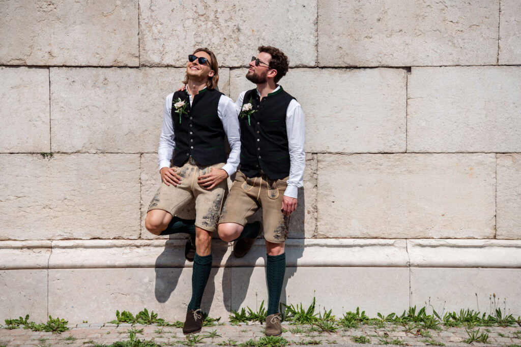 Zwei coole und schwule Männer posieren nach ihrer Hochzeit an der Bavaria in München für ihre Brautpaarportraits - stilecht mit Lederhosen und Sonnenbrille