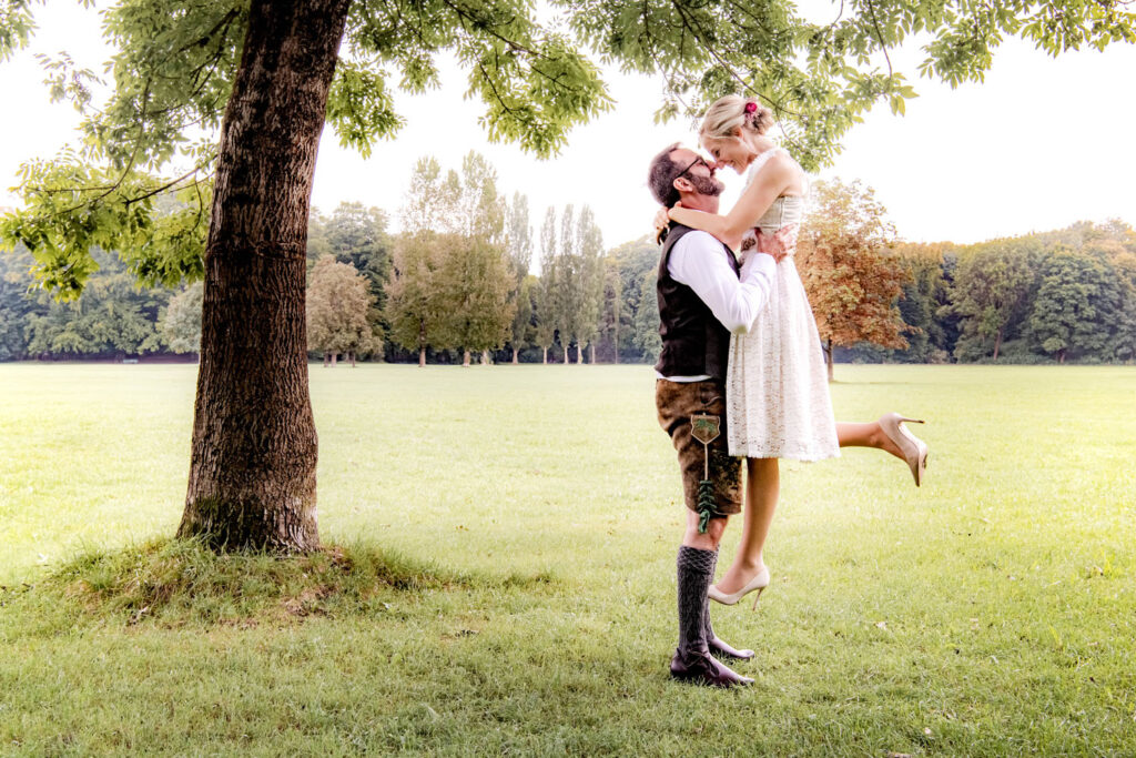 Der stolze Bräutigam hebt seine Braut hoch, beide lachen sich an - ein eindrucksvolles Hochzeitsfoto im Englischen Garten in München