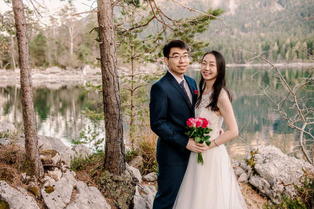 Das chinesische Brautpaar lacht beim Brautpaarshooting am Eibsee in die Kamera der Hochzeitsfotografin