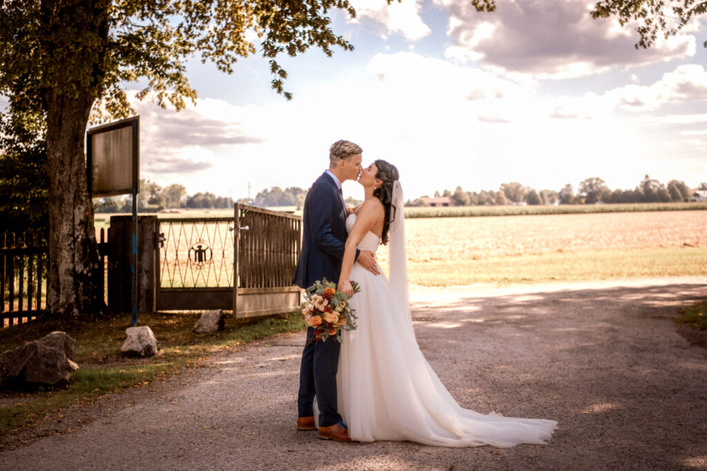 Das Brautpaar küsst sich am nach der Hochzeit Eingang der Location Waldhaus zur alten Tram bei Grünwald. Im Hintergrund ist ein grünes Feld und viel blauer Himmel zu sehen. Das Gegenlicht und der Halbschatten dieses perfekten Sommertages in München machen das Bild für die Hochzeitsfotografin perfekt.